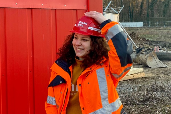 Junge Bauleiterin steht lächelnd vor einem roten Baucontainer und hält ihren Helm fest