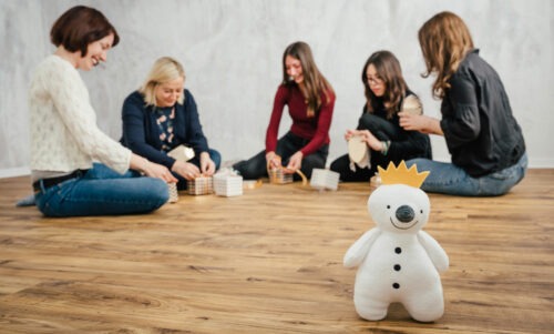 Im Hintergrund sitzen fünf Frauen auf dem Boden und basteln Geschenkverpackungen. Im Vordergrund steht ein kleiner Plüsch-Schneekönig.
